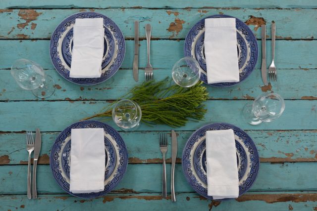 Overhead view of a rustic table setting featuring blue plates, white napkins, and silver cutlery on a weathered wooden plank. Greenery is placed in the center as a simple centerpiece. Ideal for use in articles or advertisements related to dining, home decor, rustic weddings, or farmhouse style events.