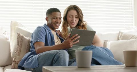 Interracial couple sharing tablet on bright sofa enjoying cozy home moment