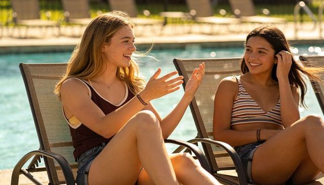Teenage friends chatting and laughing on poolside lounge chairs in sunny summer afternoon
