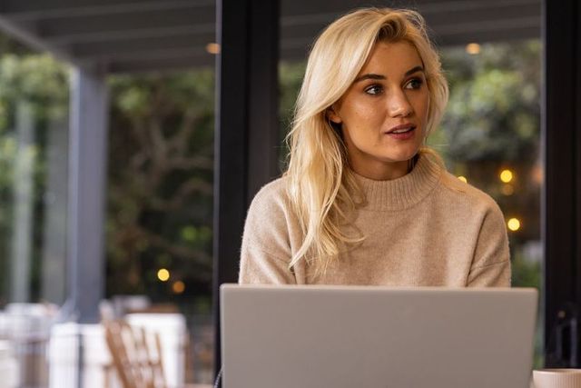 Blonde woman working remotely on laptop in cozy beige sweater with warm cafe ambience
