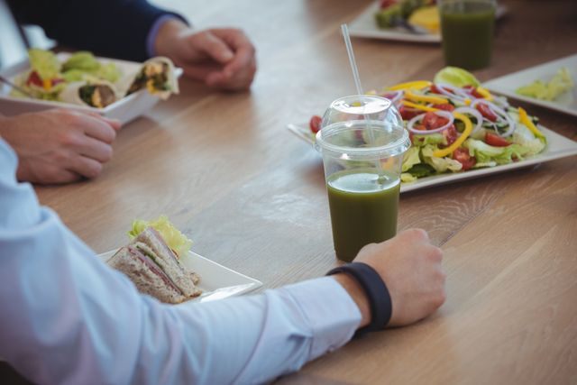 Business colleagues are having breakfast together at an office cafeteria. The table is set with healthy food options including salads, sandwiches, and green juice. This image can be used to depict corporate culture, teamwork, and healthy eating habits in a professional setting.