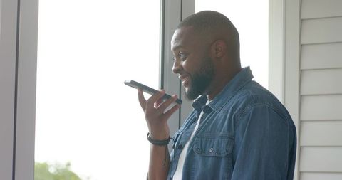 African american man using voice assistant on smartphone by glass door in casual denim