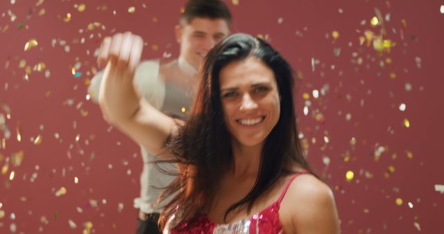 Front view close up of a happy young Caucasian woman wearing an evening dress blowing golden confetti from her hands at the camera and dancing under falling confetti at a party, a young Caucasian man dancing behind her, against a pink background