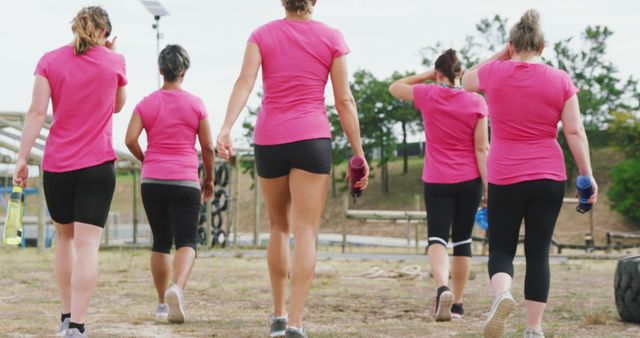 Group of women wearing pink athletic shirts and black shorts walking together after a workout outdoors in nature. They are carrying water bottles and showing camaraderie. Suitable for themes related to exercise, fitness, health, community events, and outdoor activities.