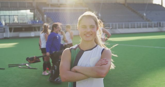 A young female field hockey player is standing on a green field, smiling confidently with arms crossed. Other players in the background are gathering with their equipment. The sun is shining, creating a warm light flare. This image can be used for promoting sporting events, health and fitness campaigns, team sports activities, and youth sports programs.