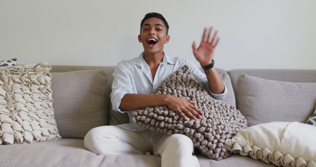 A cheerful man is relaxing on a cozy sofa, holding a textured cushion and waving happily. He is dressed in casual attire, creating a welcoming and friendly atmosphere. This can be used in lifestyle blogs, advertisements for furniture or home decor, or websites promoting relaxation and home comfort.