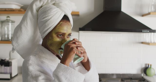 Woman wearing towel and facemask in kitchen, sipping morning coffee. Perfect for illustrating morning self-care routines, relaxation, daily skincare practices, or home comfort. Can be used in blogs, articles, advertisements on beauty and wellness, or social media campaigns promoting relaxation and self-care.