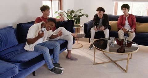 Multiracial young men playing video games on deep blue sofas in bright modern living room