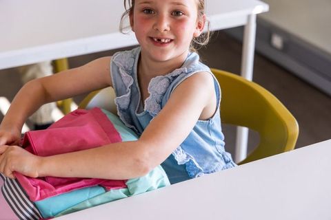 Smiling elementary school girl packing colorful pink and mint backpack at classroom desk