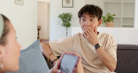 Surprised young man covering mouth while looking at smartphone on modern living room sofa