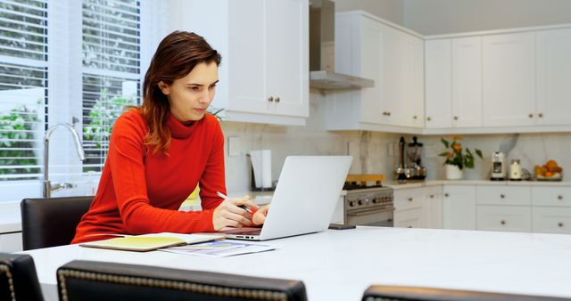 Woman in red sweater working on laptop at kitchen table. Home office setup in modern kitchen with white cabinets. Great for illustrating remote work, technology use, or home office environments.