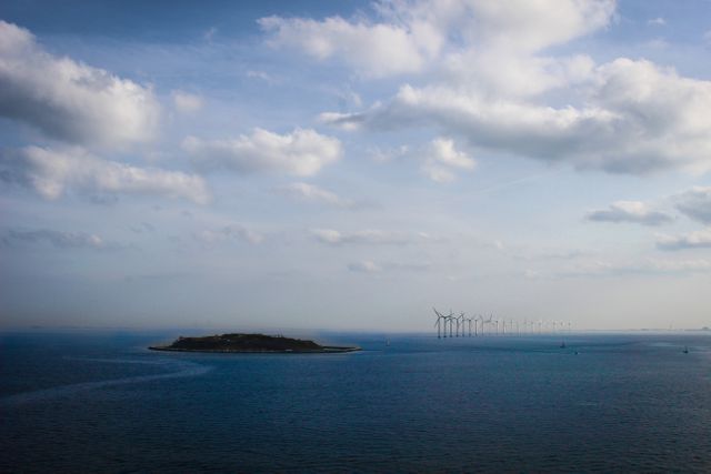 This image captures a serene sea landscape with an island in the middle of a calm ocean accompanied by wind turbines on the horizon. The sky is partly cloudy, creating a peaceful atmosphere. Perfect for illustrating concepts such as renewable energy, environmental conservation, sustainable living, and coastal scenery. Ideal for use in environmental campaigns, travel brochures, or nature magazines.