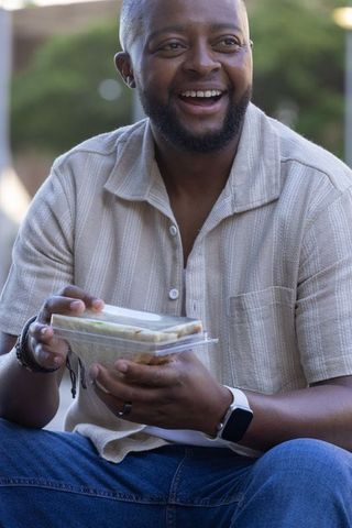 Laughing african american man holding takeout sandwich on bench outdoor lunch lifestyle