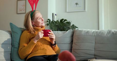 Woman wearing antler headband relaxing with coffee on cozy sofa