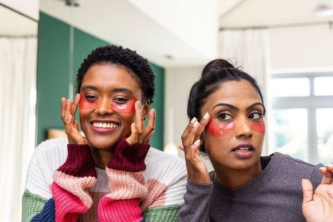 Smiling women applying red hydrogel under-eye patches while relaxing at home