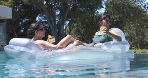 Two women relaxing on clear inflatable lounger in sunlit backyard pool holding cocktails