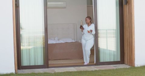 African american woman sipping coffee and checking smartphone in loungewear at sliding door
