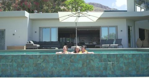 Three women relaxing in turquoise infinity pool with cocktails under patio umbrella