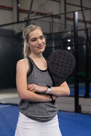 Fit female padel player standing on blue indoor court holding black racket and neon ball