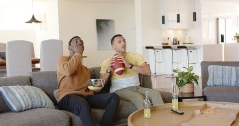 Diverse male friends watching football at home on modern sofa with chips pizza drinks