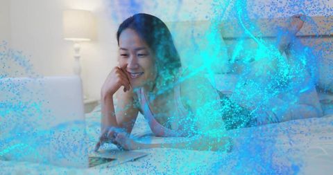 Korean woman relaxing on bed, smiling while using laptop with blue particle overlay