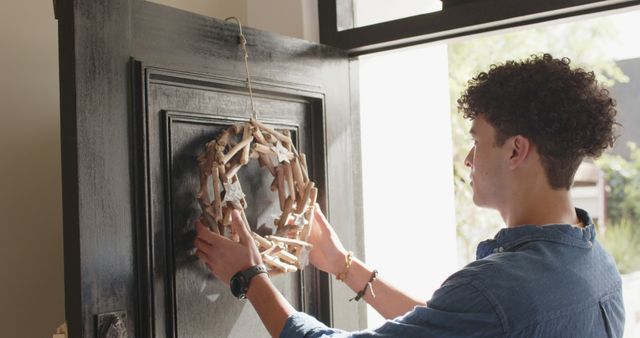 Young man hanging a rustic driftwood wreath on a black front door, focusing on the concept of home decor and welcoming guests. Useful for articles on DIY decor, seasonal decorations, and front door styling tips.