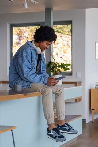 African american man sitting on kitchen island with smartphone, denim jacket and headset