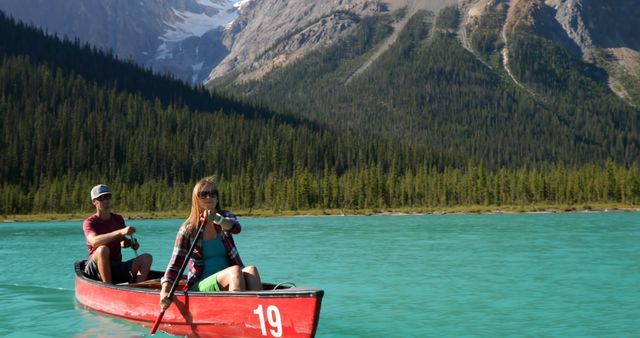 Couple paddling their red canoe on a clear turquoise lake surrounded by majestic mountains and dense forest. Perfect for promoting travel, outdoor adventures, and nature activities. Use for articles, travel guides, and advertising scenic destinations.