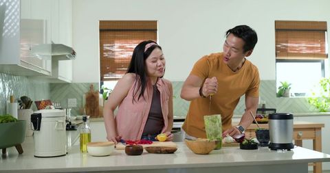 Asian couple preparing fresh green smoothie together on sunlit modern kitchen island