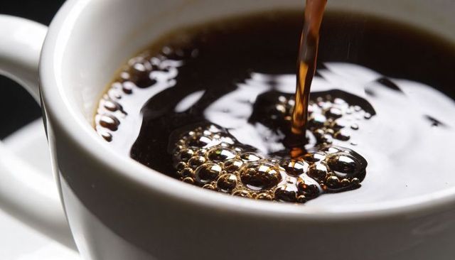 Pouring dark brewed coffee into white ceramic cup with reflective bubbles closeup macro