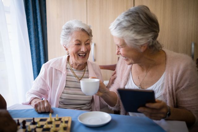 Two senior women are enjoying coffee and playing chess at a nursing home. They are laughing and engaging in a friendly conversation, showcasing the joy of companionship and leisure activities in a retirement setting. This image can be used for promoting elderly care services, retirement homes, social activities for seniors, and the importance of friendship and socializing among the elderly.