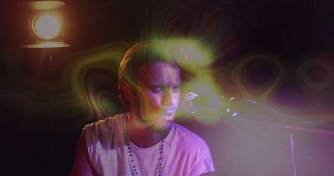 Singing fair-skinned male vocalist performing under psychedelic neon spotlight with mic