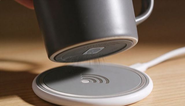 Matte gray ceramic mug landing on wireless charging pad with crumbs on wooden desk