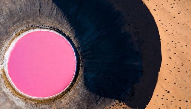 Aerial view of circular pink salt lake contrasting with dark desert shadow and white crust
