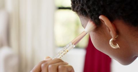 African american woman applying makeup with brush by sunlit window, closeup beauty selfcare