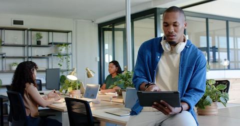 Man using tablet with headphones in bright coworking space while colleagues collaborating