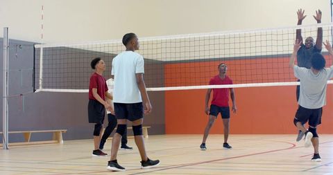Diverse male teens playing competitive indoor volleyball, jumping and blocking at net on court