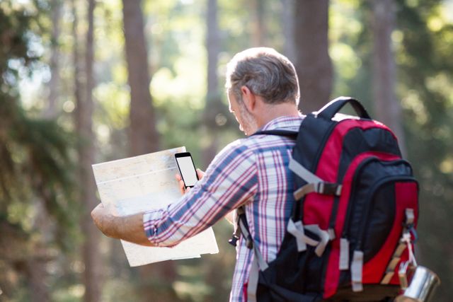 Senior male hiker using a map and smartphone for navigation in a forest. Ideal for content related to outdoor adventures, travel, navigation technology, active lifestyles, and nature exploration.