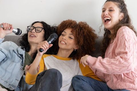 Diverse women singing karaoke on sofa, casual cozy living room music night together