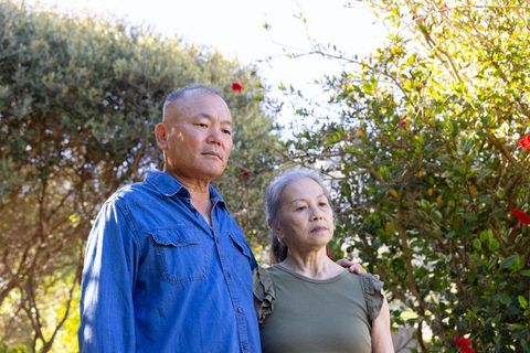 Senior asian couple standing in sunlit garden resting hand on partner quiet companionship