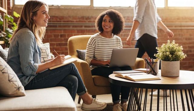 Two women collaborating in sunlit loft coworking space using laptop and notebook