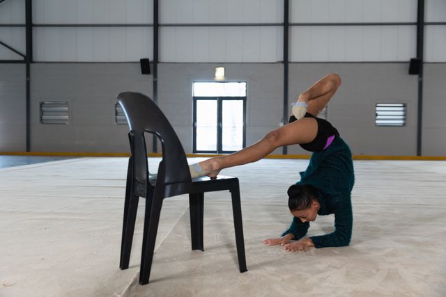 Biracial female gymnast practicing at the gym, doing a handstand with a chair, stretching her legs and warming up. Gymnast training hard for competition.