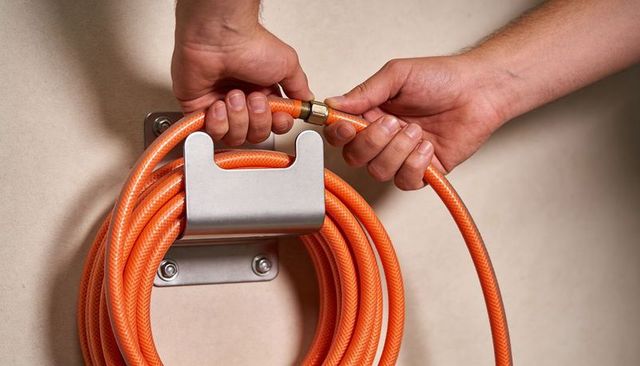Hands connecting orange garden hose to wall-mount reel with brass coupling, closeup
