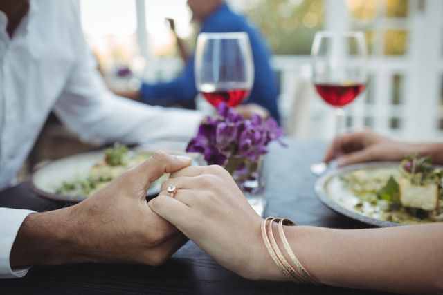 Couple holding hands across a restaurant table, enjoying a romantic meal with wine. Ideal for use in content related to romance, relationships, dining experiences, special occasions, and engagement announcements.