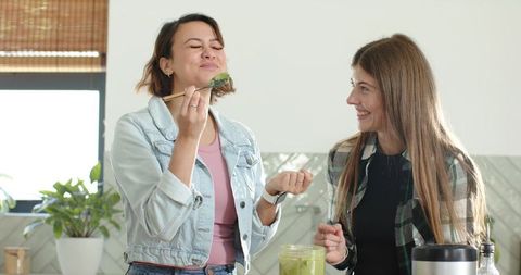 Diverse friends tasting green smoothie in sunlit kitchen smiling, enjoying healthy drink