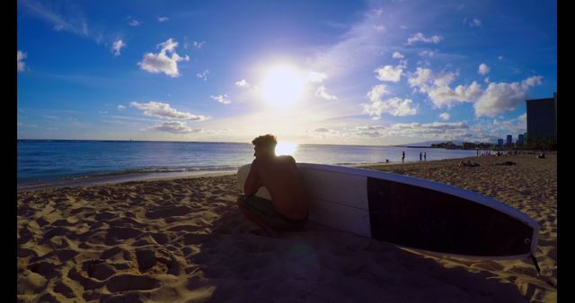 This image shows a man sitting on a sandy beach next to a surfboard while watching the sunset over the ocean. Ideal for use in travel blogs, vacation promotional materials, surfing lifestyle content, and web design featuring beach and ocean themes.