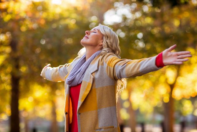 Young woman standing with arms outstretched, enjoying the autumn season in a park. Ideal for use in lifestyle blogs, wellness articles, advertisements for outdoor activities, or promotions for autumn fashion.