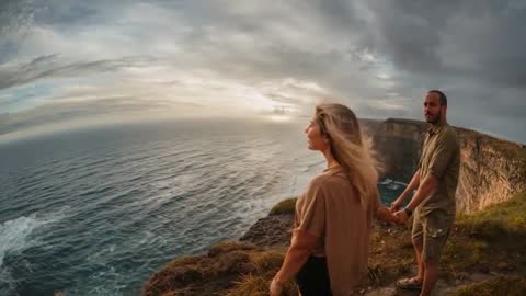 Couple holding hands watching ocean sunset from windswept cliff edge at golden hour