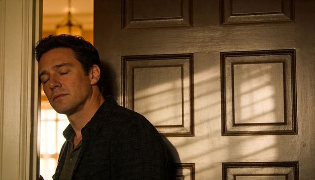 Man leaning and resting against doorframe in warm backlit hallway, contemplative mood