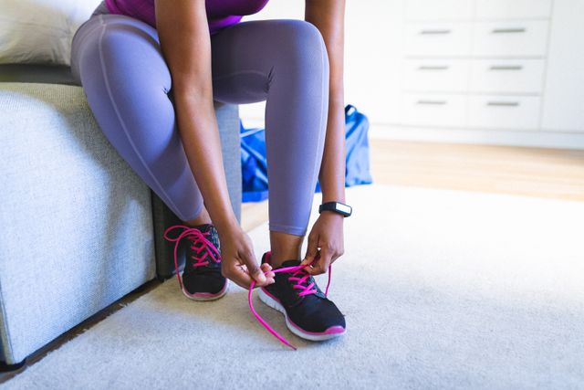 This image shows an African American woman tying her shoelaces before a workout at home. It is perfect for use in fitness blogs, healthy lifestyle promotions, workout routines, and articles about home exercise. The casual and relatable setting makes it ideal for content focused on everyday fitness and wellbeing.
